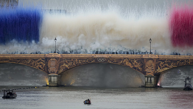 The Olympic torch travels by boat as ceremonial smoke in the colors of the France flag appear over the Seine River Paris, France, during the opening ceremony of the 2024 Summer Olympics, Friday, July 26, 2024. (AP Photo/Matthias Schrader)