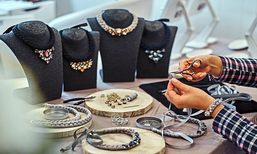 Elegantly dressed woman makes handmade necklaces, working with needles and thread in jewelry workshop.